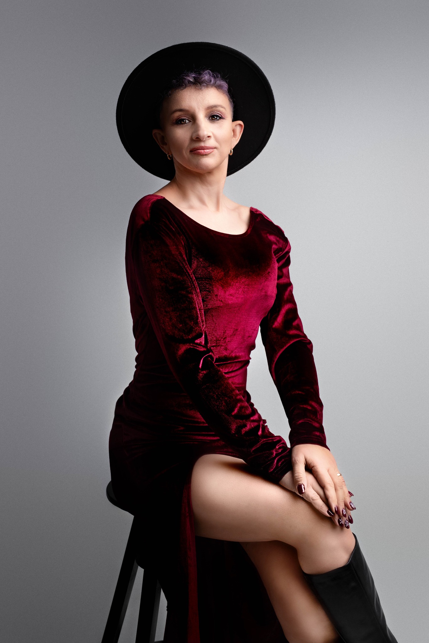 Portret kobiety siedzącej na krześle, ubrana w bordową, welurową sukienkę z rozcięciem, czarny kapelusz i czarne kozaki, z fioletowymi włosami i manicure w odcieniach bordo.