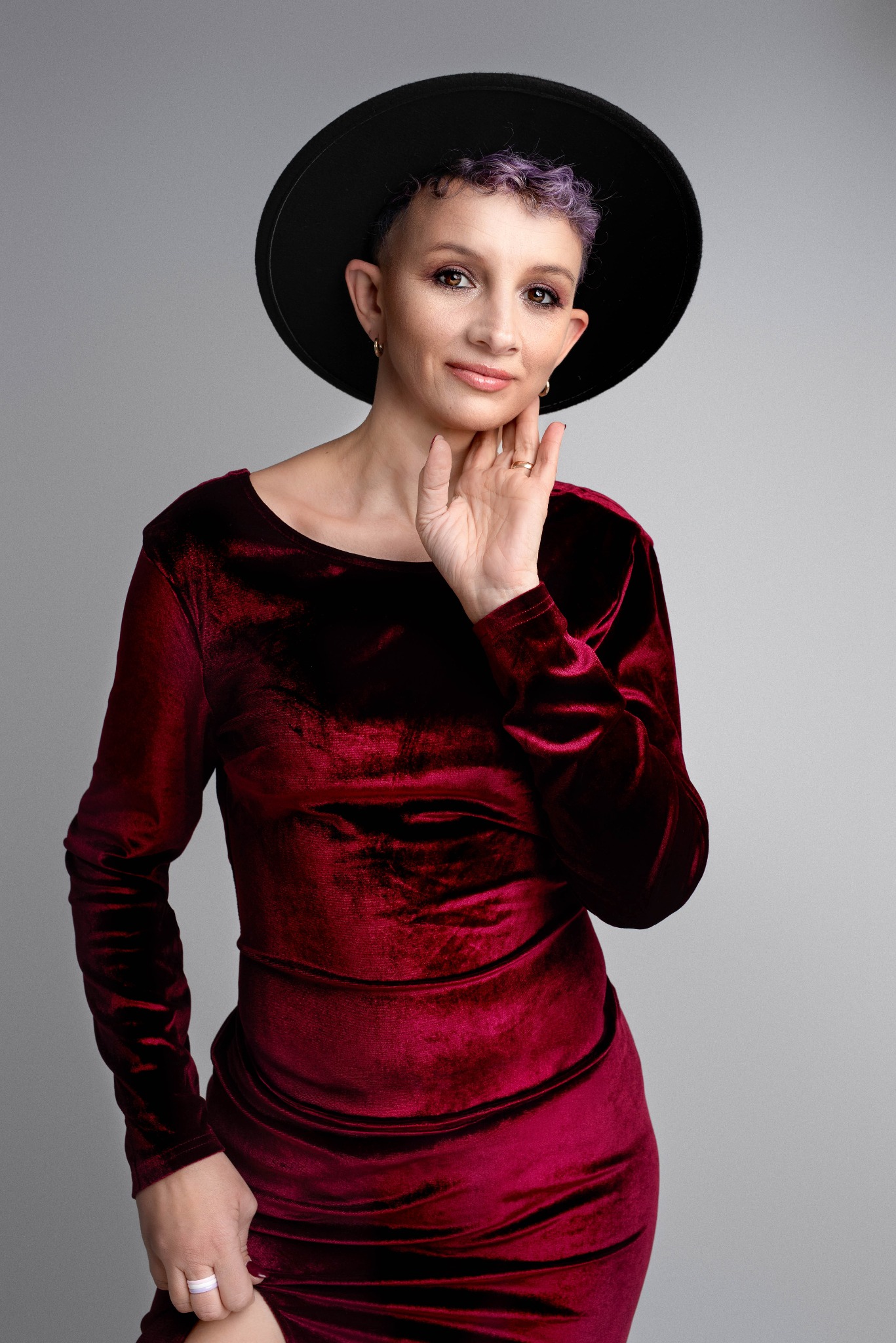 Portret kobiety w bordowej welurowej sukience i czarnym kapeluszu, z krótkimi fioletowymi włosami, dłonią dotykającej policzka, pozującej na szarym tle.