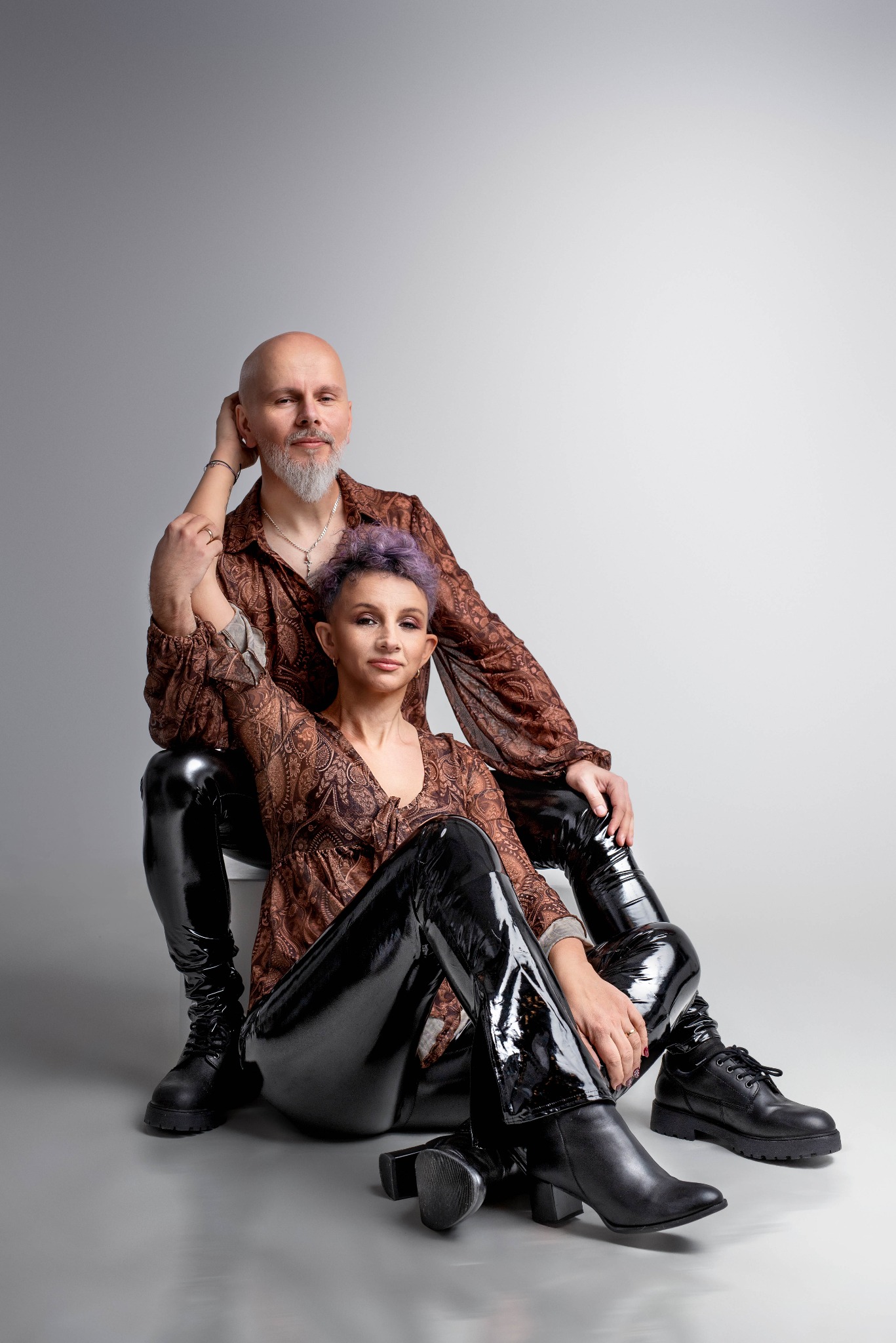 Portret pary: mężczyzna z siwą brodą opiera rękę na ramieniu kobiety z krótkimi fioletowymi włosami, oboje w brązowych wzorzystych koszulach i czarnych lakierowanych spodniach, siedzą na białym tle.