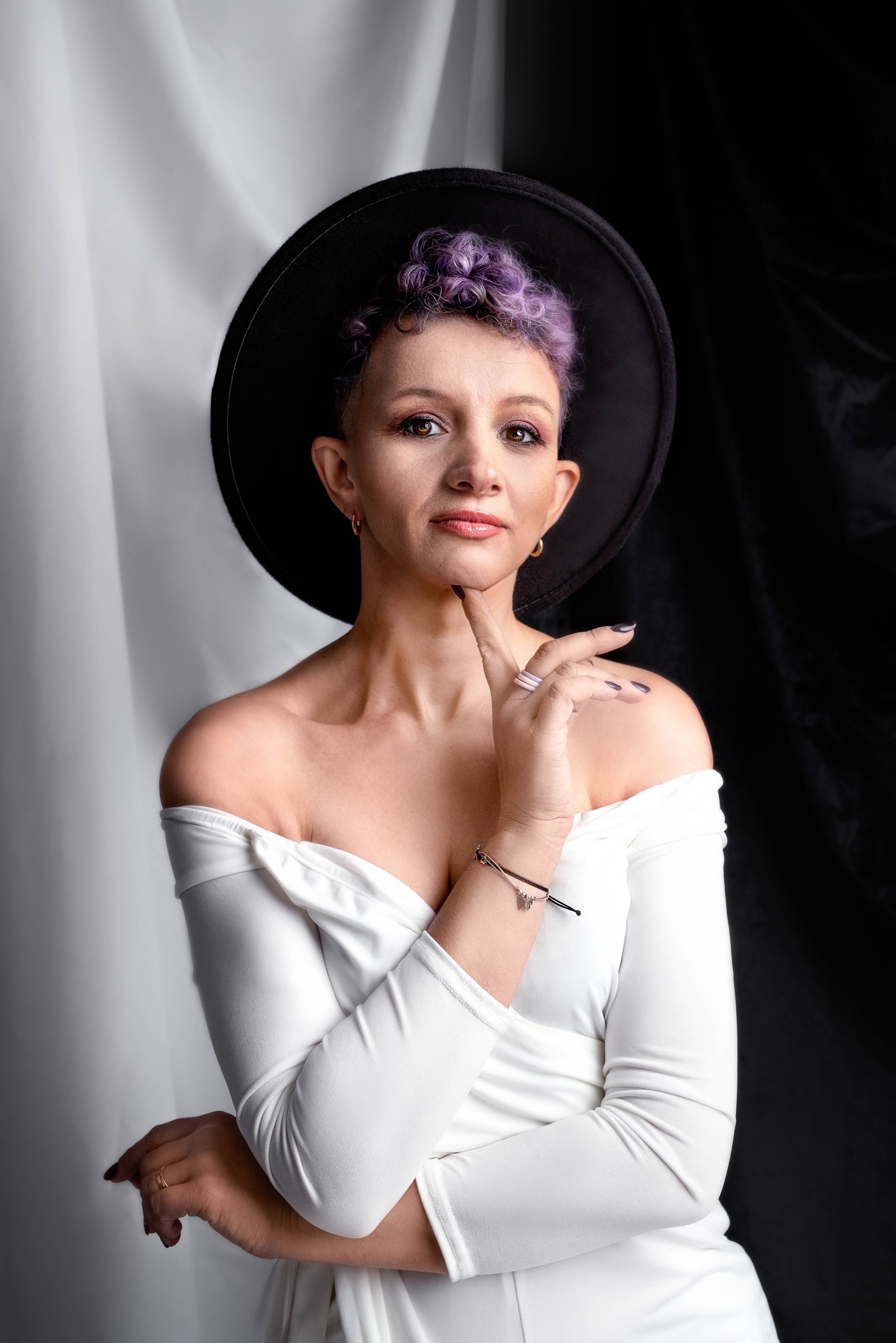 Portret kobiety z fioletowymi włosami w czarnym kapeluszu, ubrana w białą sukienkę z odkrytymi ramionami, pozująca na tle białej i czarnej tkaniny.