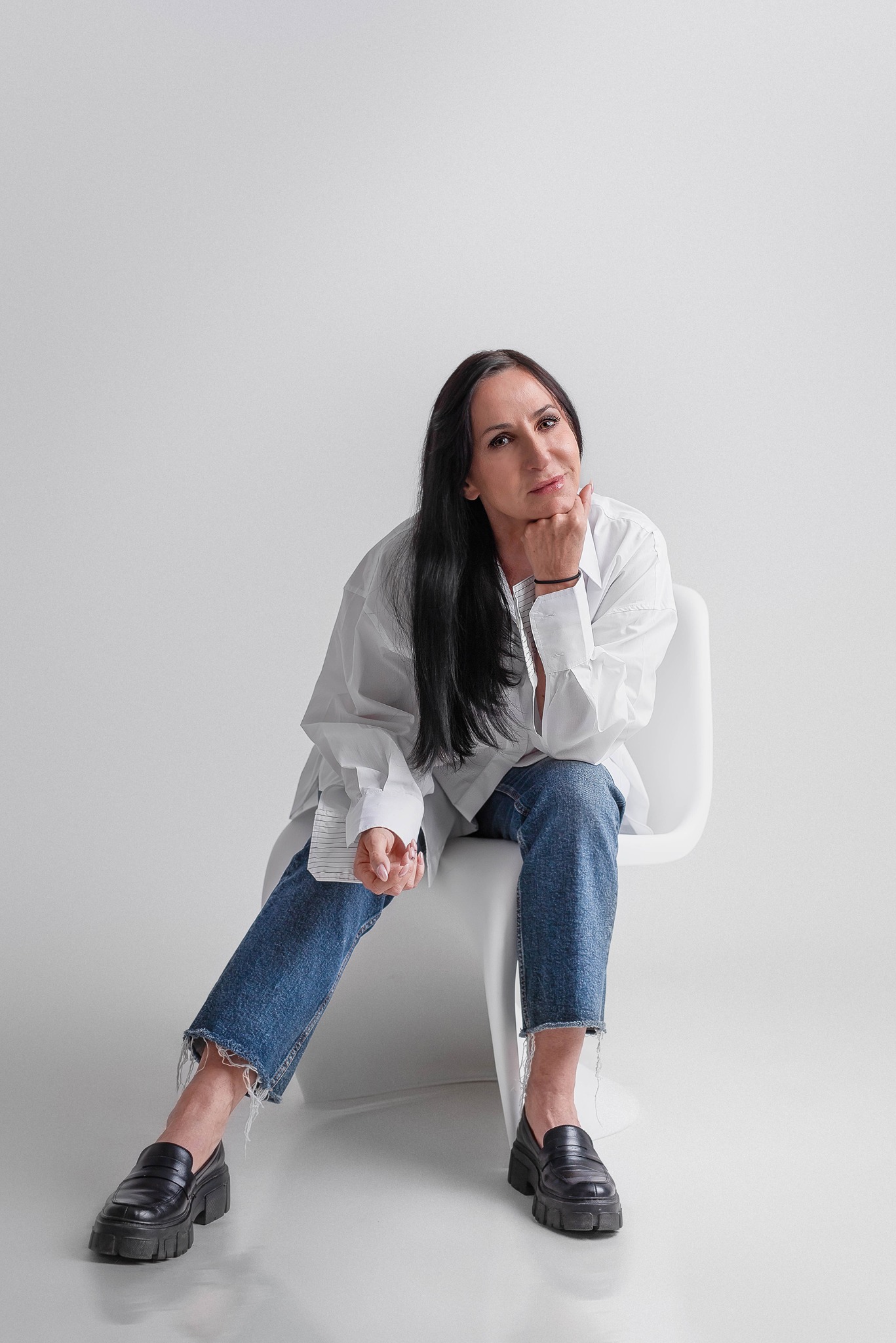 Portret kobiety siedzącej na białym krześle w obszernym białym męskim koszuli i dżinsach, z ręką podpartą pod brodą, na jasnoszarym tle.