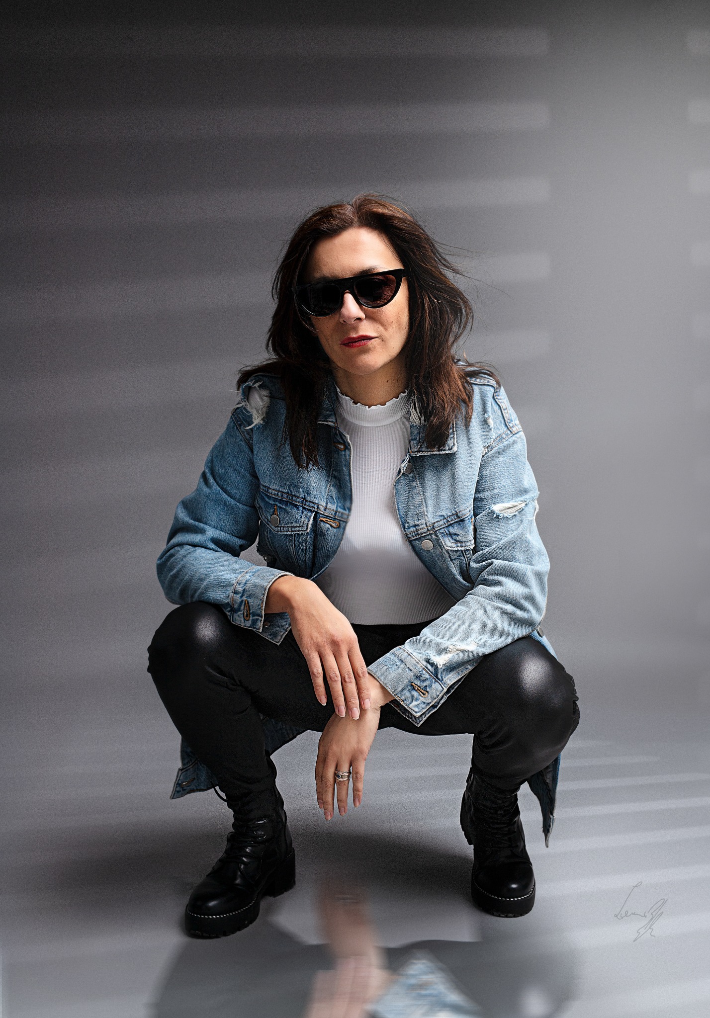 Kobieta w dżinsowej kurtce i skórzanych spodniach kuca na szarym tle, nosi ciemne okulary i ma pierścionek na palcu.