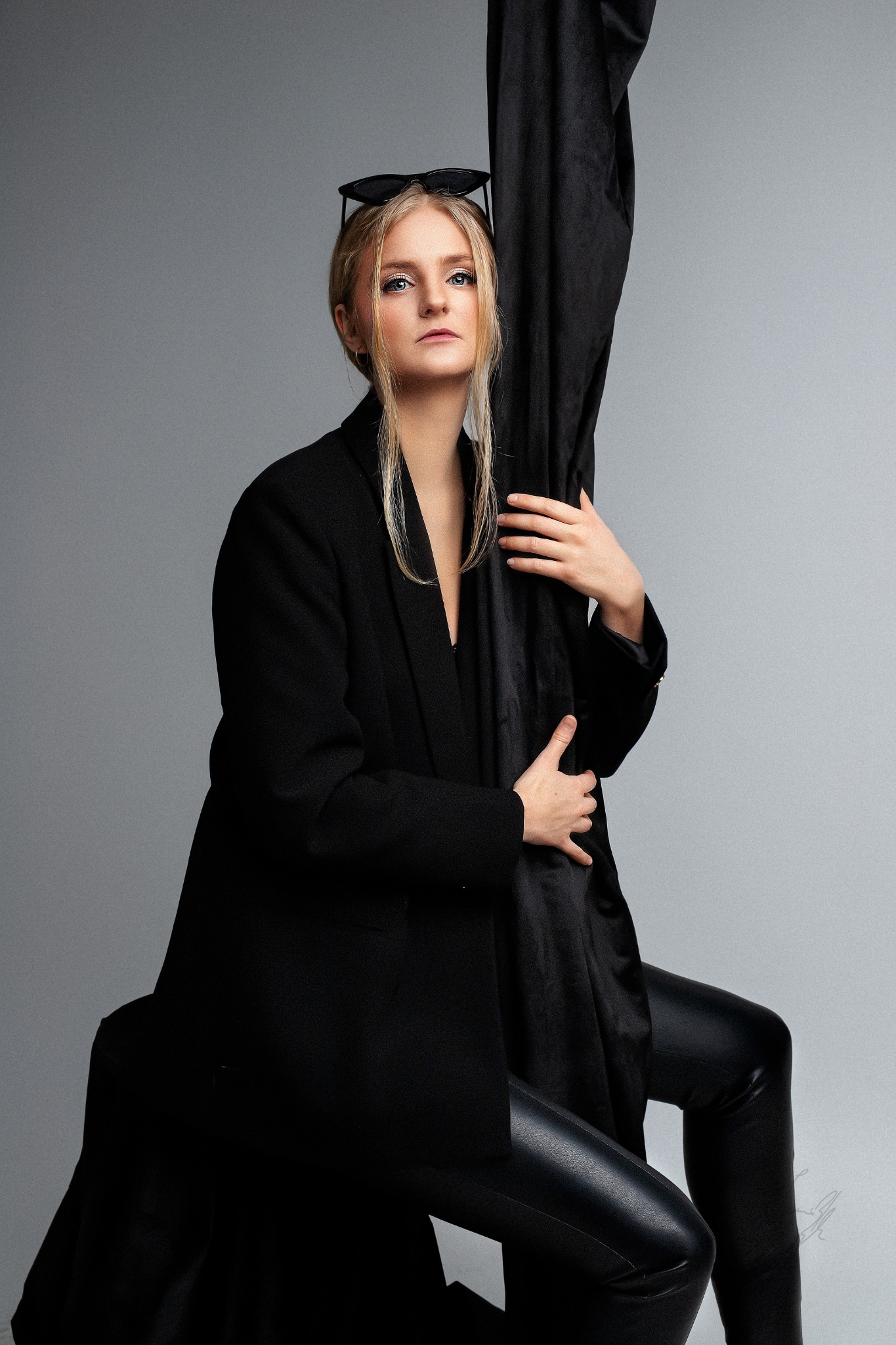 Portret kobiety w czarnym garniturze i skórzanych spodniach, pozującej na szarym tle z czarnym materiałem, okulary na głowie.