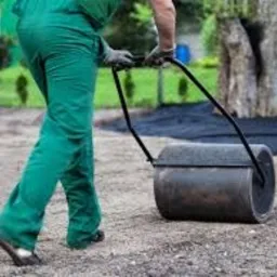 Pracownik w zielonym kombinezonie ubija ziemię walcem ręcznym na przygotowanym podłożu z agrowłókniny, widoczny trawnik i drzewo w tle.