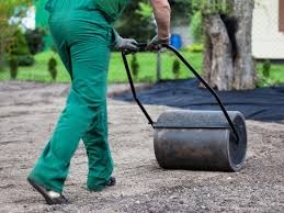 Pracownik w zielonym kombinezonie ubija ziemię walcem ręcznym na przygotowanym podłożu z agrowłókniny, widoczny trawnik i drzewo w tle.