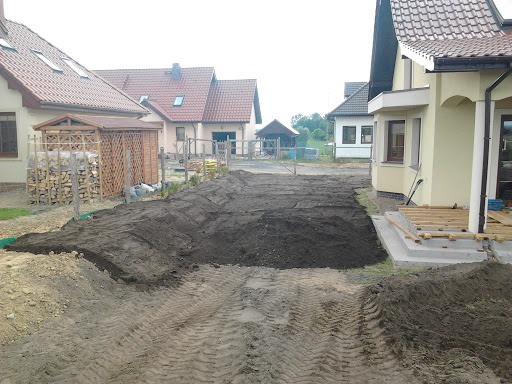 Przygotowany teren pod budowę lub ogród, wyrównana ziemia z widocznymi śladami opon, w tle zabudowa jednorodzinna i drewutnia z zapasem drewna.