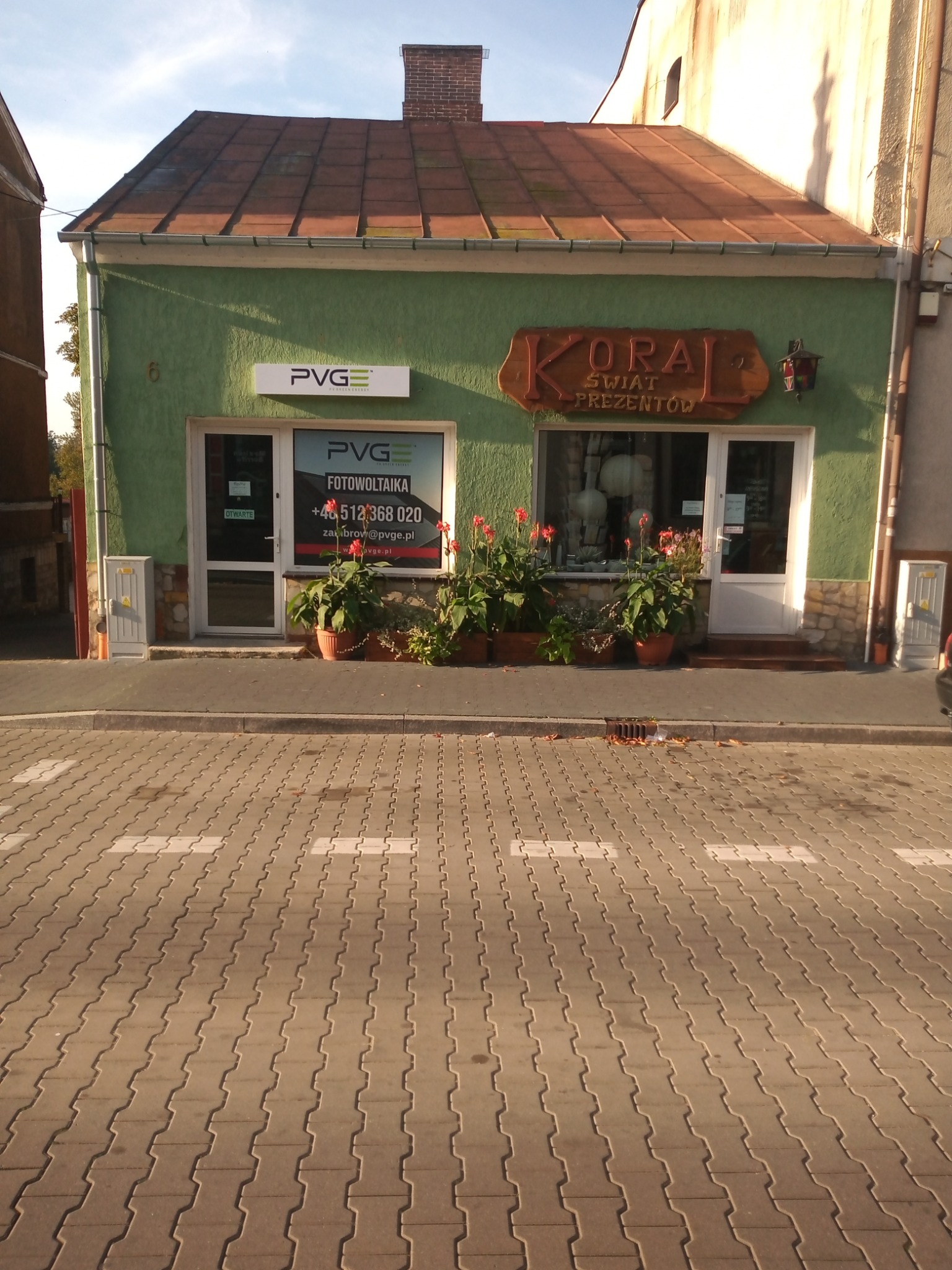 Fasada budynku z dwoma lokalami: PVG Fotowoltaika z oknem z naklejonymi danymi kontaktowymi i KORA Świat Prezentów, oba lokale oddzielone kwiatami w donicach.