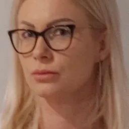 Portret kobiety z blond włosami w okularach z brązowymi oprawkami, patrzącej wprost, z delikatnym makijażem i subtelną biżuterią.