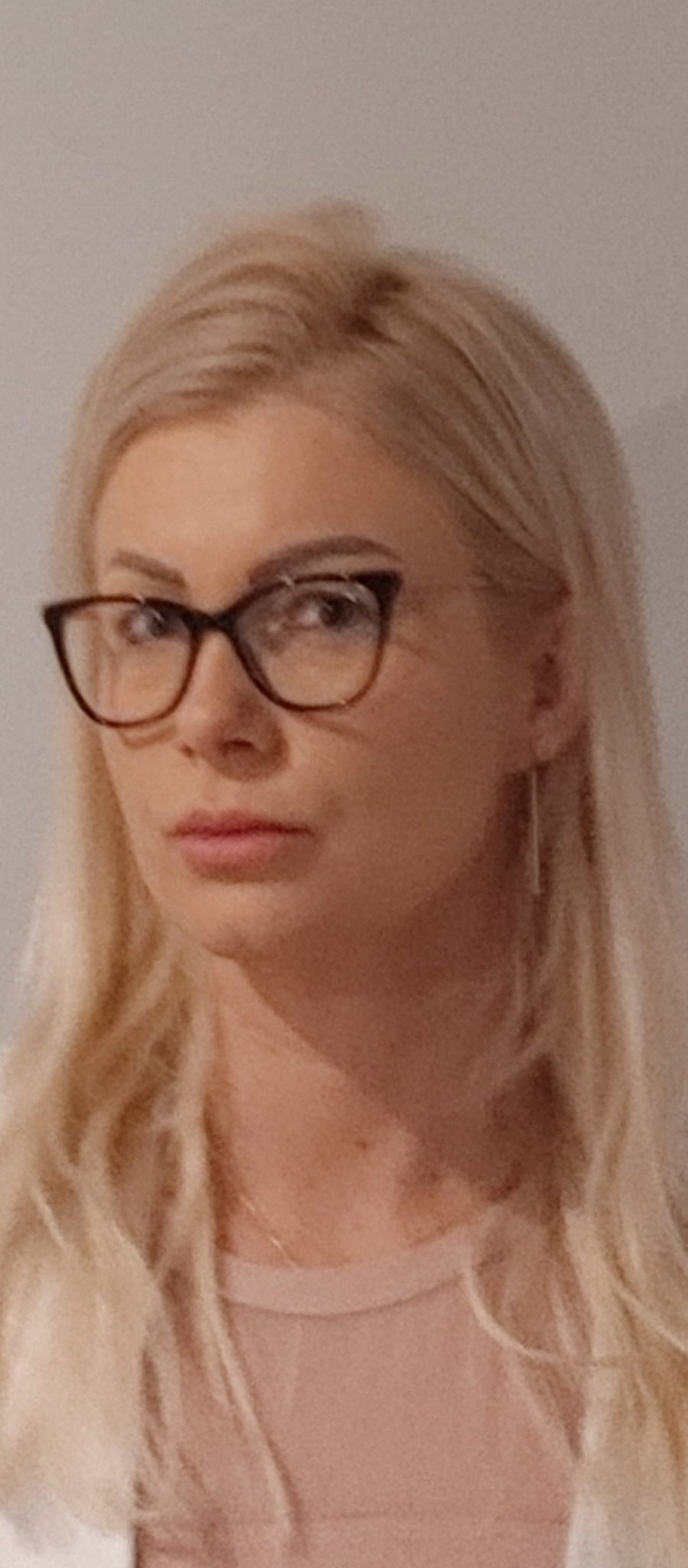 Portret kobiety z blond włosami w okularach z brązowymi oprawkami, patrzącej wprost, z delikatnym makijażem i subtelną biżuterią.