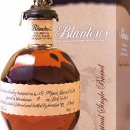 Butelka bourbonu Blanton's Original Single Barrel z metalową figurką jeźdźca na koniu na korku, stojąca obok pudełka z tym samym napisem.