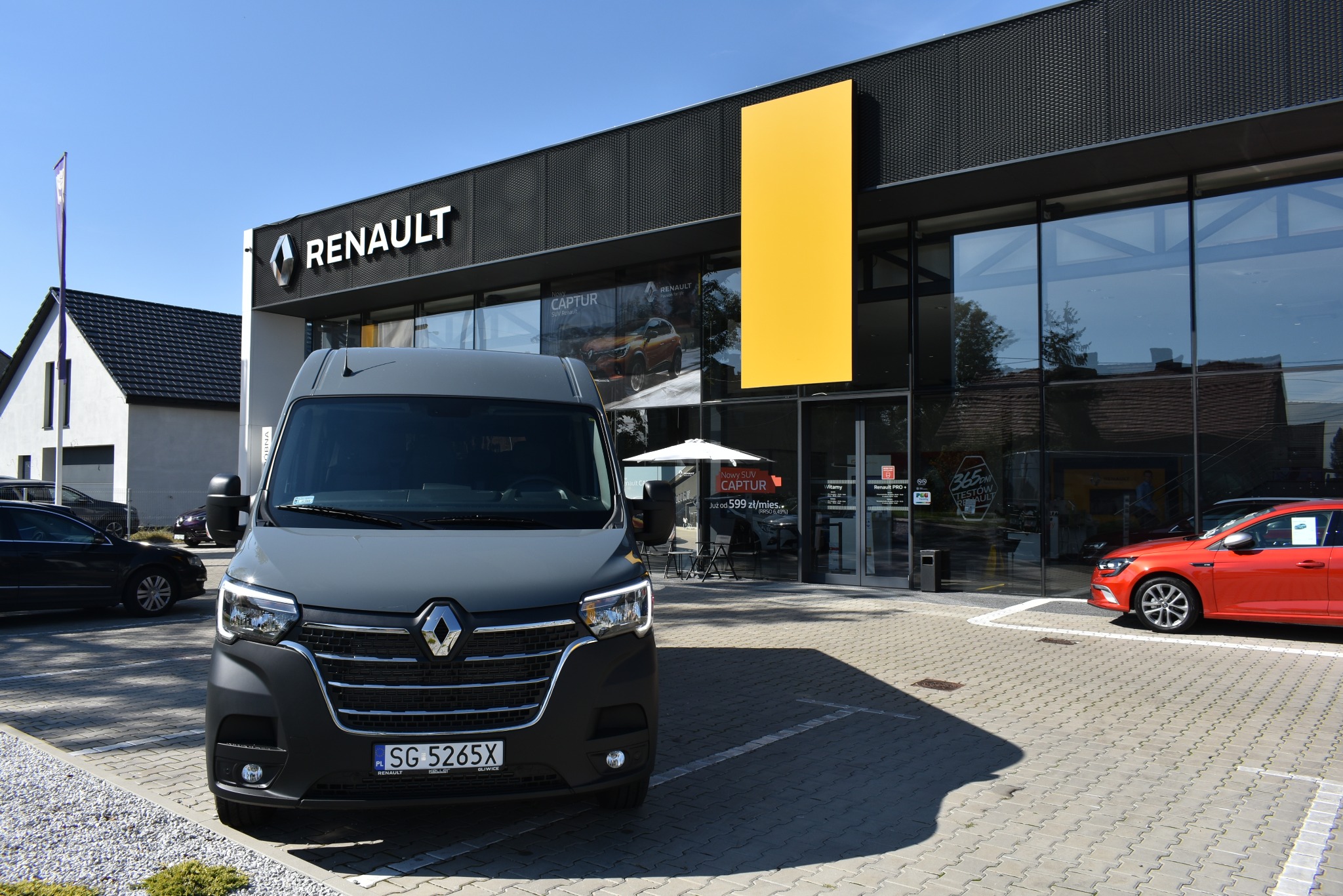 Szary bus Renault Master przed salonem samochodowym Renault, z widocznym logo firmy i ekspozycją nowych modeli Captur w tle.