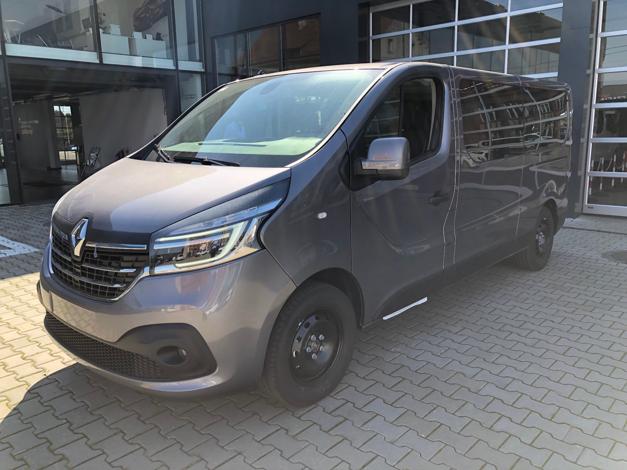 Szary Renault Trafic z czarnymi felgami stalowymi na kostce brukowej przed salonem samochodowym z przeszklonymi drzwiami garażowymi.