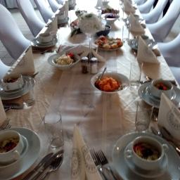 Fabryka Smaków Legionowo - Długi stół bankietowy z białym obrusem, nakryty dla wielu gości, z białymi krzesłami w tle, dekorowany kwiatami i różnorodnymi potrawami w białych naczyniach. Na pierwszym planie widoczne...