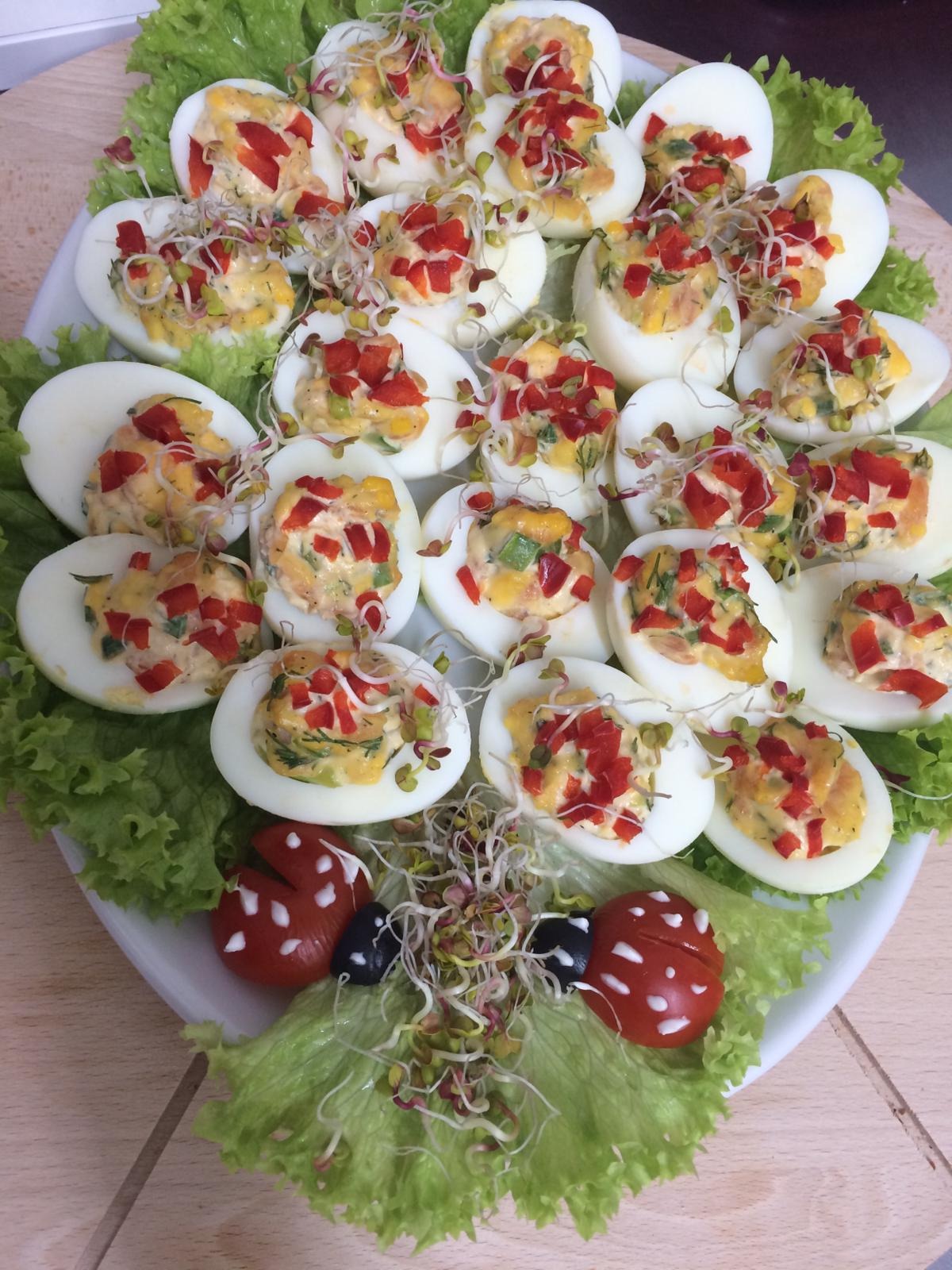 Talerz z jajkami faszerowanymi, udekorowanymi czerwoną papryką i kiełkami, ułożonymi na liściach sałaty z dekoracjami w kształcie biedronek z pomidorów i oliwek.