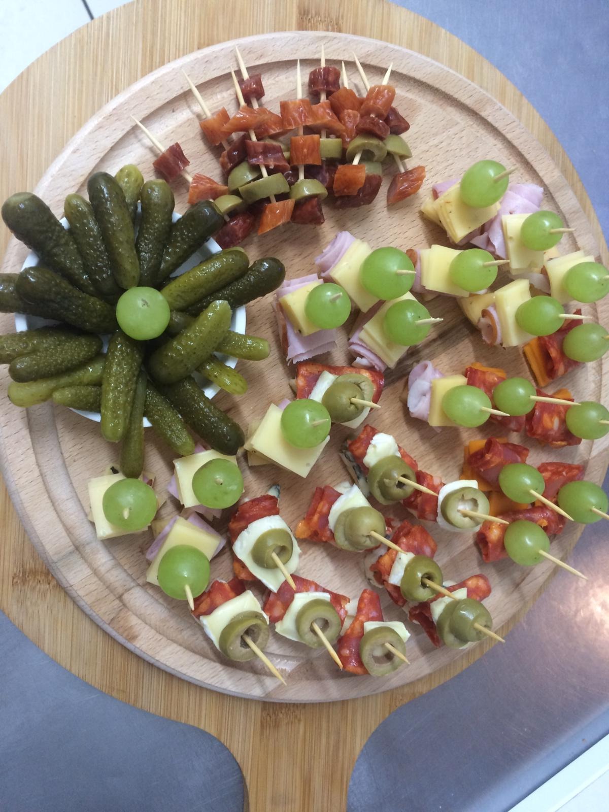 Drewniana deska z przekąskami: korniszony w białej miseczce, koreczki z salami i oliwek, koreczki z szynki, sera i winogron, koreczki z chorizo, sera i winogron, ułożone na okrągłej desce...