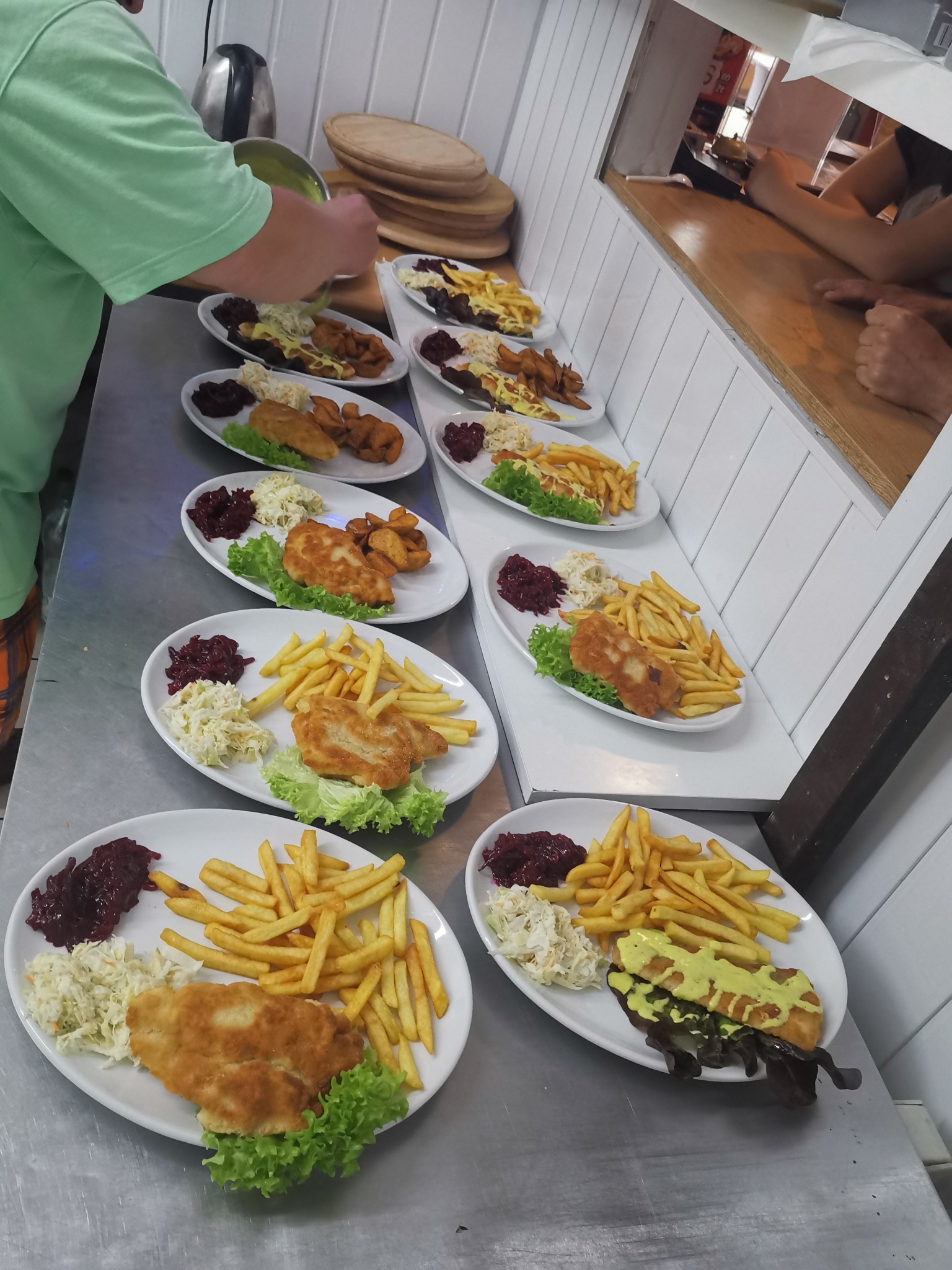 Przygotowywanie porcji obiadowych: smażona ryba z frytkami, surówką i buraczkami na białych talerzach, sos nakładany z metalowego naczynia, widok zza lady.