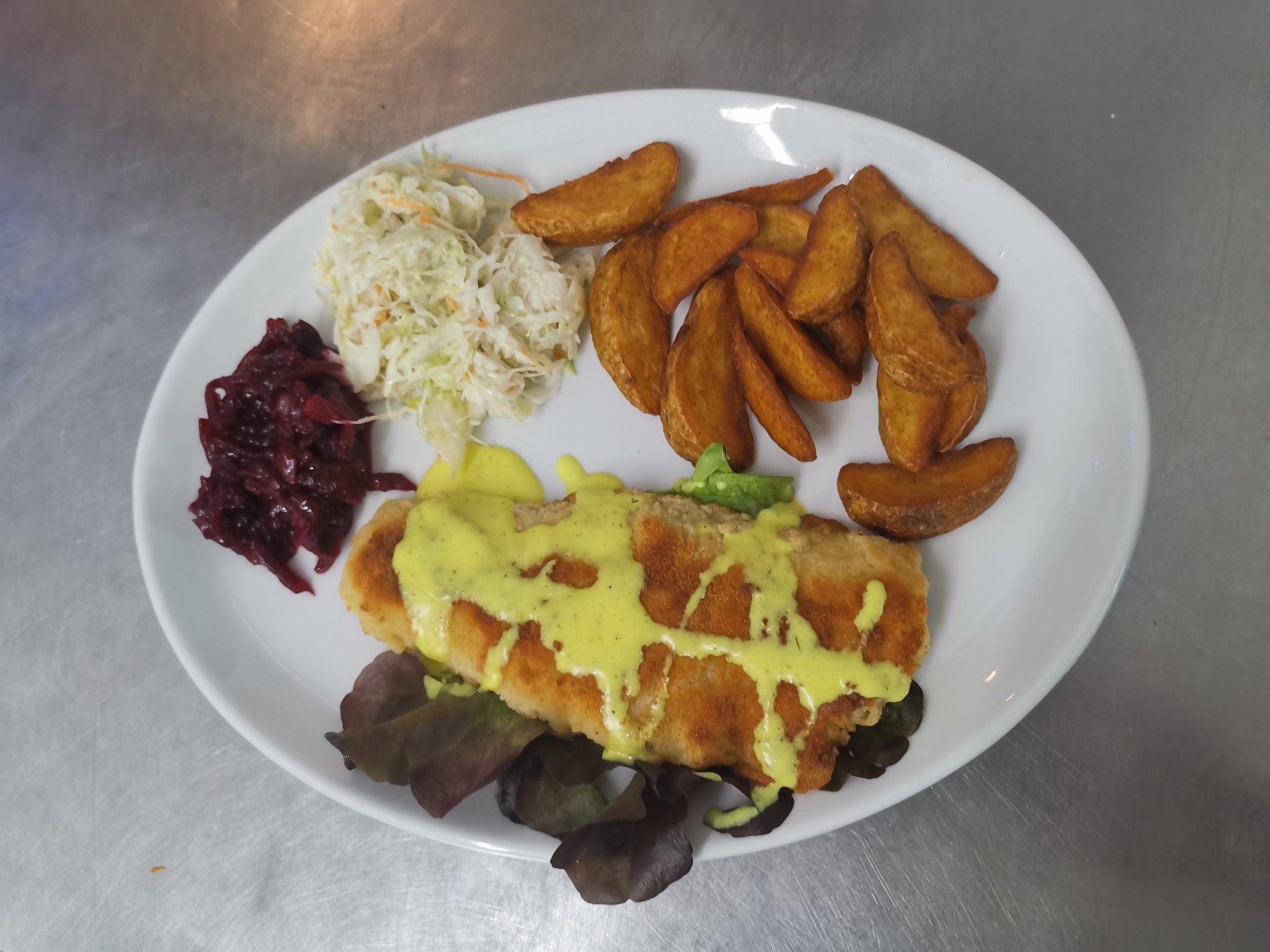 Biały talerz z panierowaną rybą polaną sosem, frytkami z ziemniaków, surówką z kapusty i buraczkami na tle metalowego blatu.