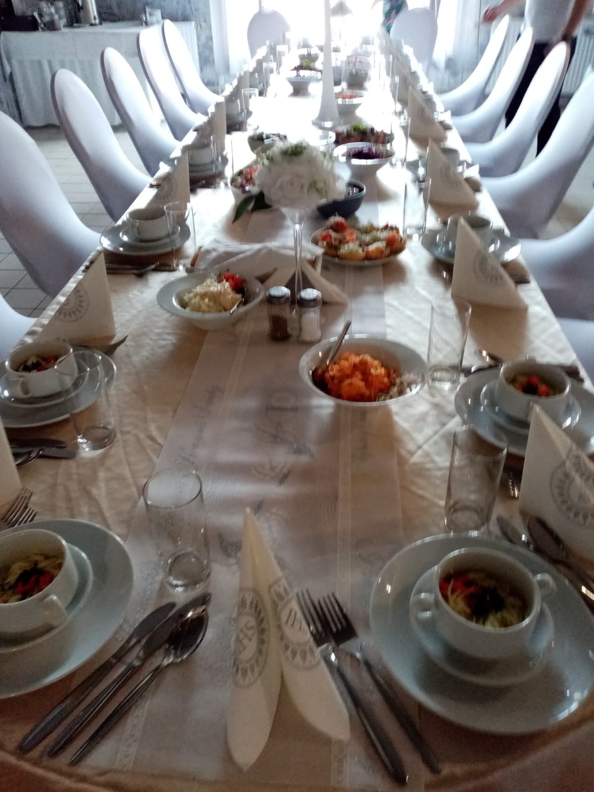 Długi stół bankietowy z białym obrusem, nakryty dla wielu gości, z białymi krzesłami w tle, dekorowany kwiatami i różnorodnymi potrawami w białych naczyniach. Na pierwszym planie widoczne...