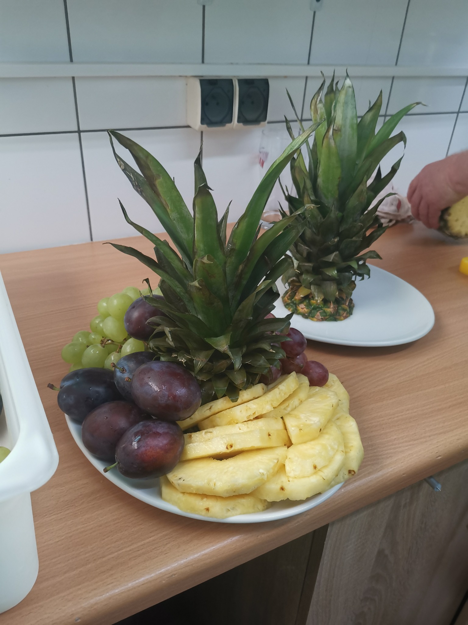 Dekoracja stołu z ananasów, śliwek i winogron na białym talerzu, przygotowana na drewnianym blacie w kuchni.