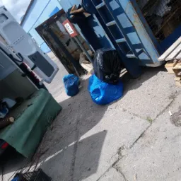 Otwarty furgon z załadowanymi gałęziami i zielonymi odpadami, obok niebieski kontener na śmieci i paleta drewniana, worki na śmieci w kolorze niebieskim i czarnym.