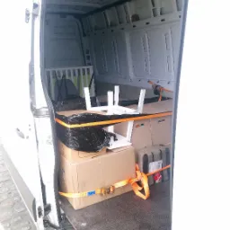 Otwarta przestrzeń ładunkowa białego furgonetki wypełniona kartonami, meblami w częściach i zabezpieczona pomarańczowymi pasami transportowymi, widok z zewnątrz.