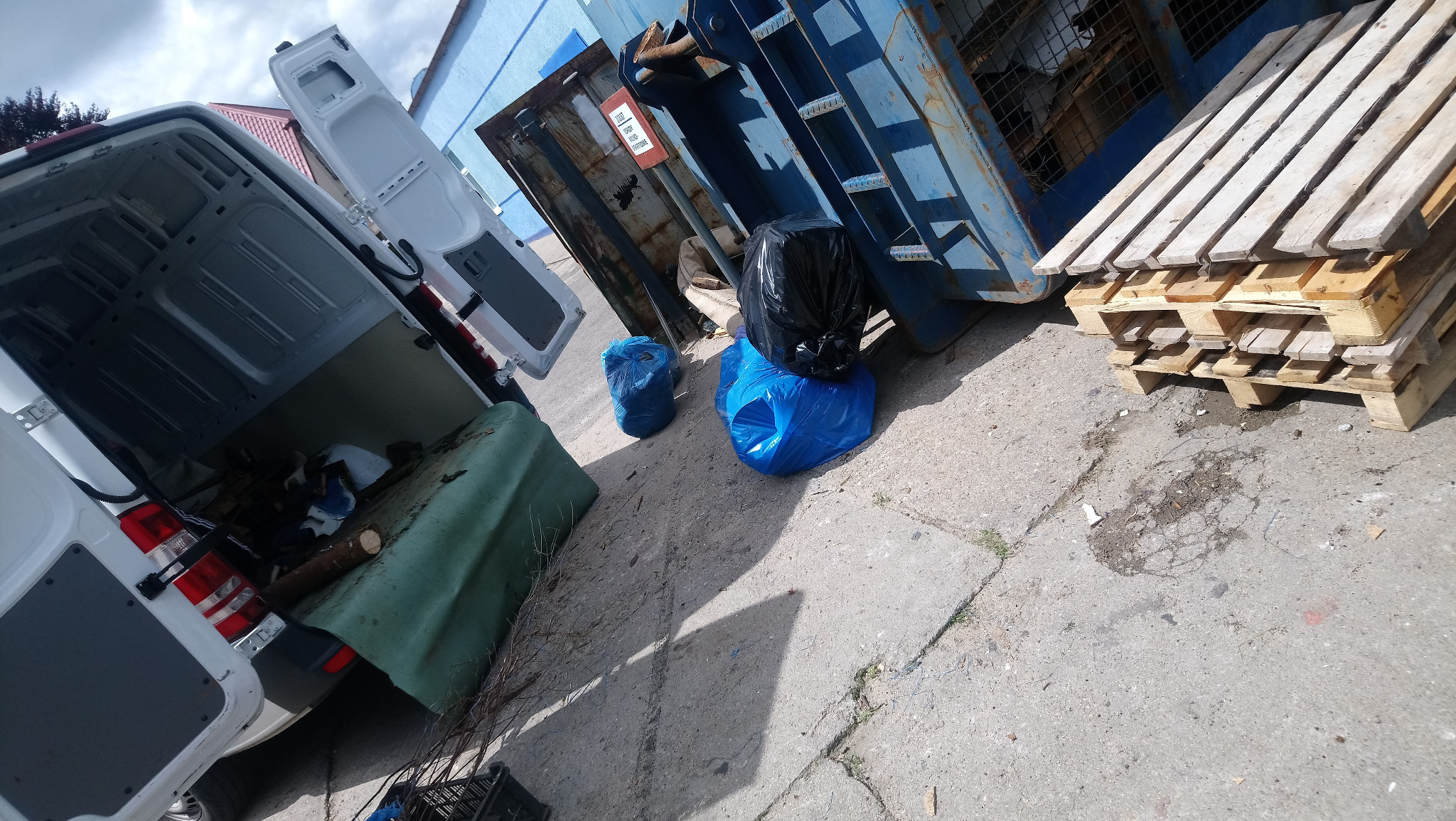 Otwarty furgon z załadowanymi gałęziami i zielonymi odpadami, obok niebieski kontener na śmieci i paleta drewniana, worki na śmieci w kolorze niebieskim i czarnym.