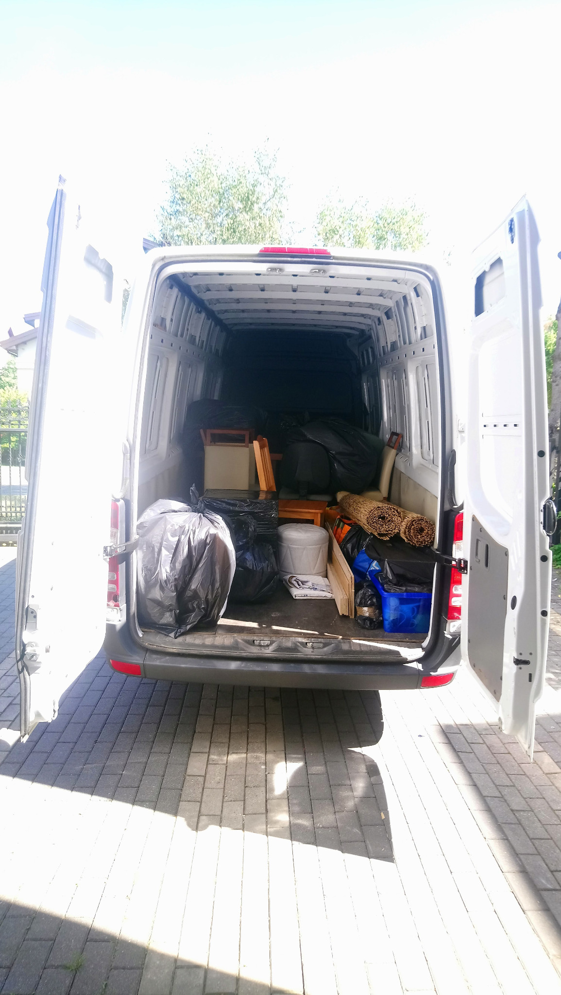 Wnętrze białego busa z otwartymi drzwiami załadowane meblami, workami i kartonami podczas dnia, widoczne z zewnątrz na tle brukowanej powierzchni.