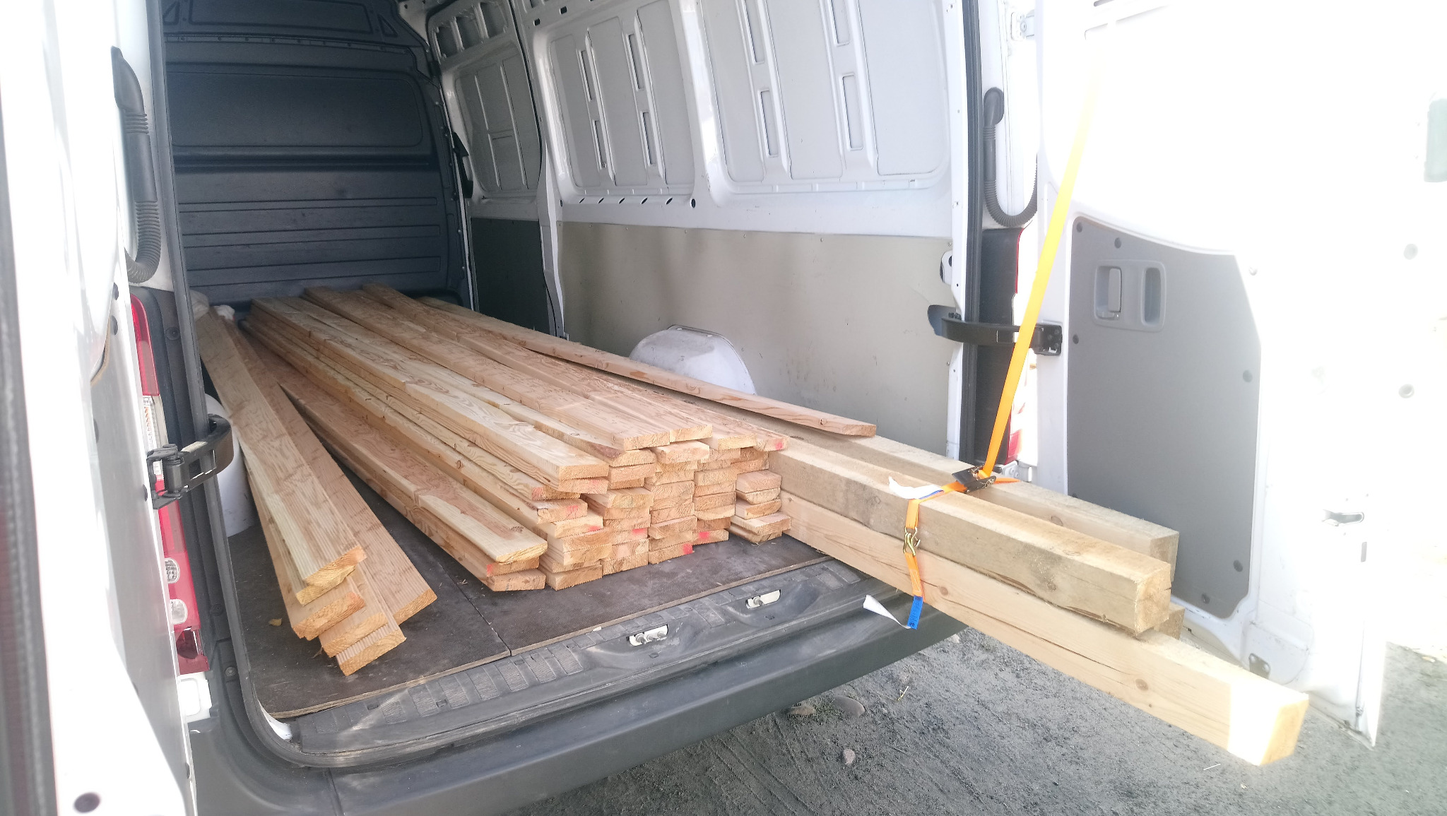 Wnętrze białego samochodu dostawczego załadowanego drewnem budowlanym, zabezpieczonym żółtymi pasami transportowymi.