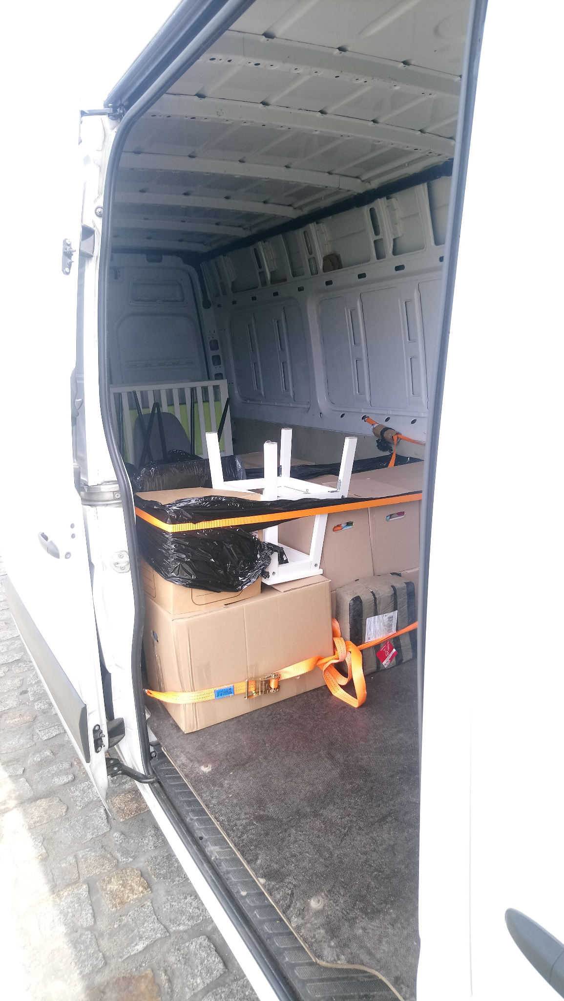 Otwarta przestrzeń ładunkowa białego furgonetki wypełniona kartonami, meblami w częściach i zabezpieczona pomarańczowymi pasami transportowymi, widok z zewnątrz.