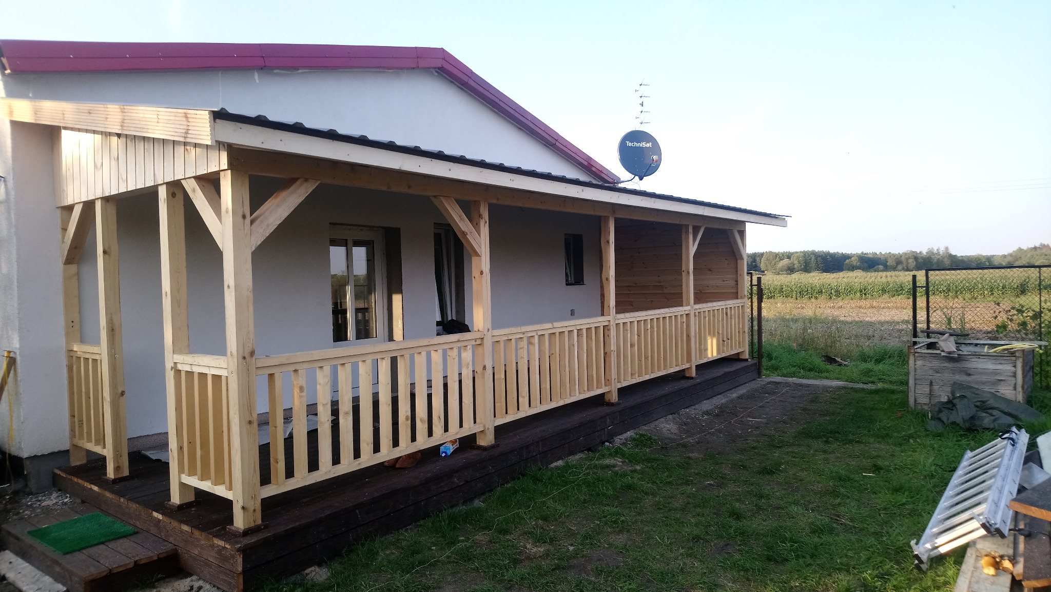 Surowa drewniana konstrukcja zadaszonego tarasu przy domu z widoczną anteną satelitarną, w tle pole kukurydzy i drabina oparta o trawę.