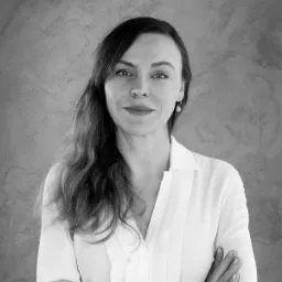 Portret kobiety w białej koszuli z rękami skrzyżowanymi na tle szarej ściany o fakturze betonu, w czerni i bieli, widoczny kolczyk w uchu.