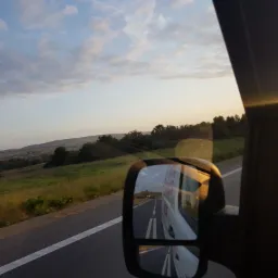 Widok z wnętrza pojazdu: krajobraz za oknem i odbicie w lusterku bocznym ukazujące jadący za nim pojazd na drodze, oświetlone promieniami zachodzącego słońca.