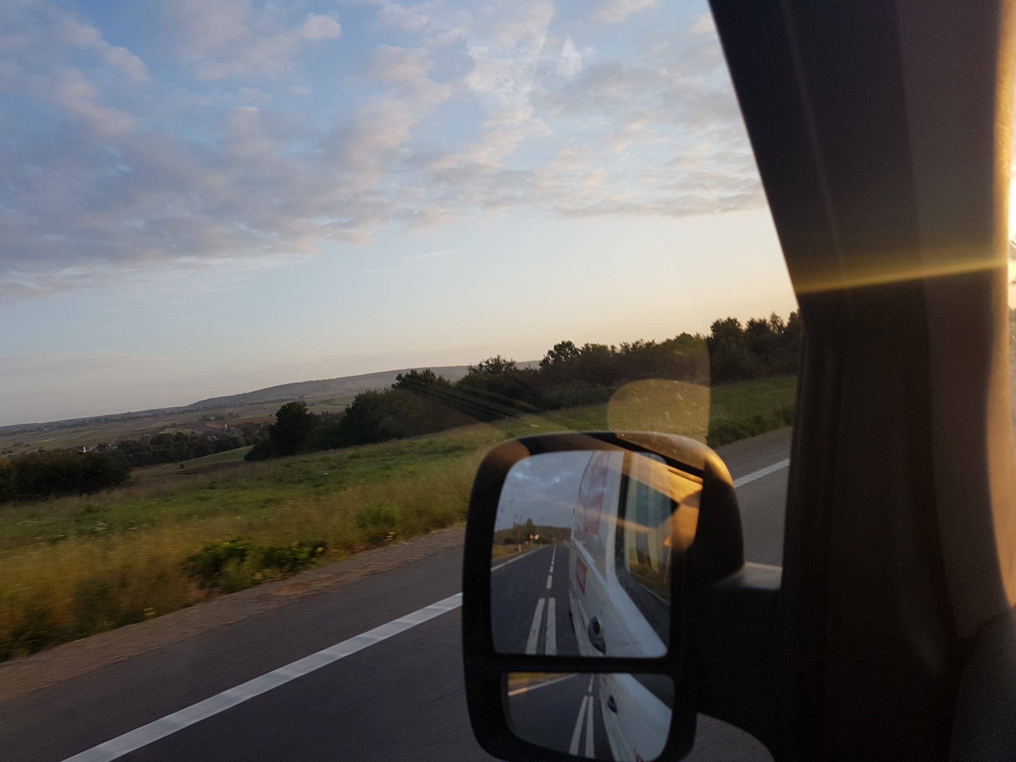 Widok z wnętrza pojazdu: krajobraz za oknem i odbicie w lusterku bocznym ukazujące jadący za nim pojazd na drodze, oświetlone promieniami zachodzącego słońca.