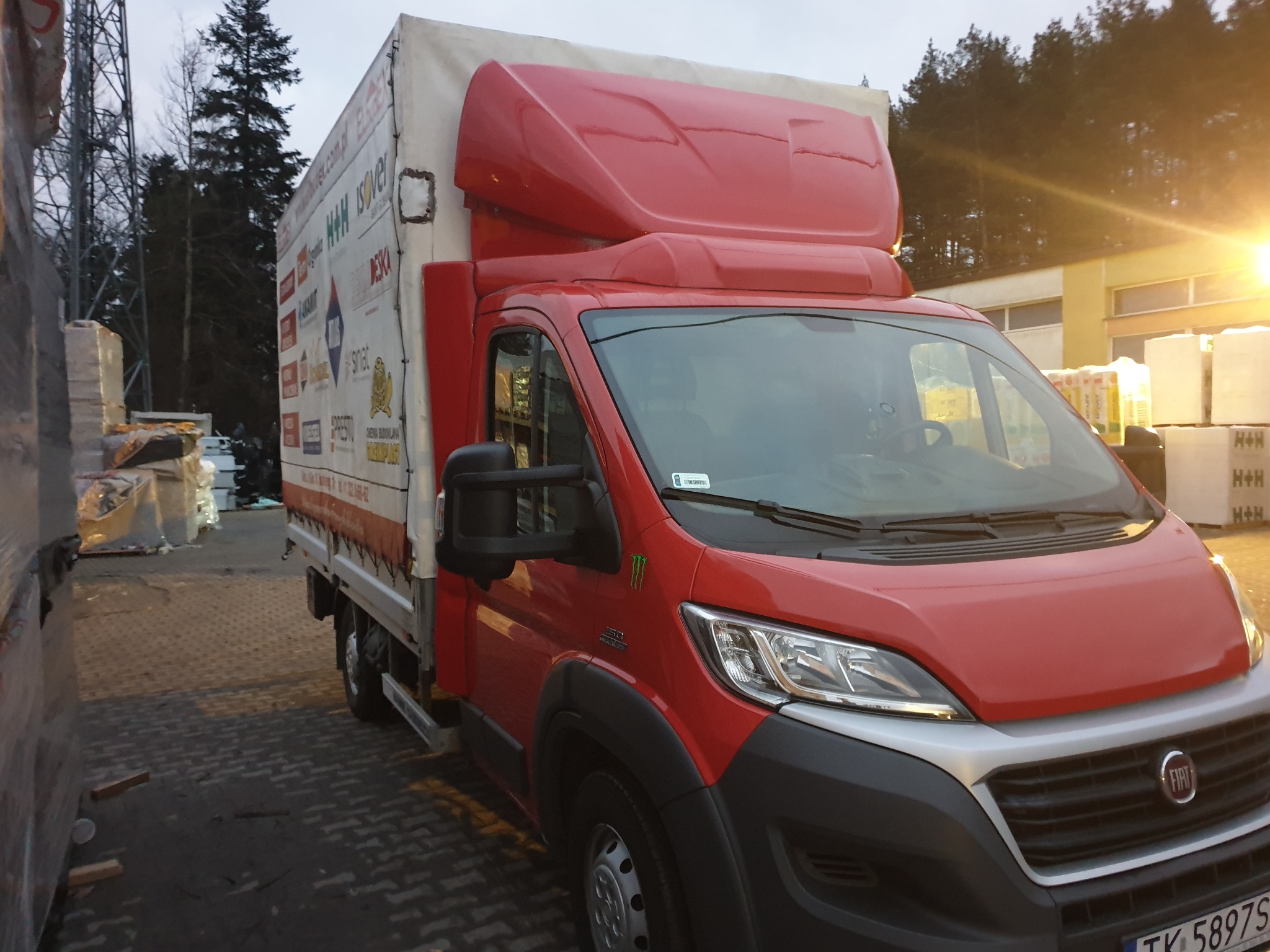 Czerwony bus dostawczy z plandeką, zaparkowany w Kielcach na placu z materiałami budowlanymi, widok z przodu.