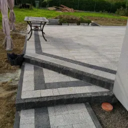 Ułożona kostka brukowa w odcieniach szarości z ciemniejszym obramowaniem, tworząca schody i taras z elementami dekoracyjnymi, widoczny stół ogrodowy i przygotowane podłoże pod trawnik.