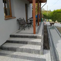 Ułożone schody i chodnik z kostki granitowej w odcieniach szarości, z widocznym fragmentem przygotowanego wykopu pod obrzeża, na tle domu z drewnianymi kolumnami i zielenią.