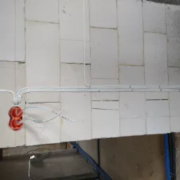 Instalacja przewodów elektrycznych w białych rurkach PCV, przytwierdzonych do ściany z bloczków, z widocznymi pomarańczowymi puszkami pod gniazdka.