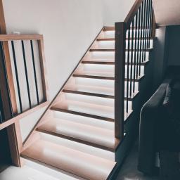 Sławomir Tytuła Usługi stolarskie - Drewniane schody z podświetleniem LED pod każdym stopniem, balustrada z drewnianymi słupkami i czarnymi, pionowymi elementami, widok od dołu, jasne płytki podłogowe.