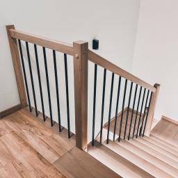 Sławomir Tytuła Usługi stolarskie - Widok z góry na drewniane schody z jasnego drewna z czarnymi metalowymi balustradami, oświetlenie ścienne w tle, widoczne fragmenty drewnianej podłogi i drzwi.