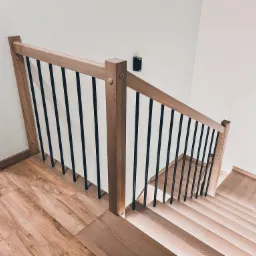 Widok z góry na drewniane schody z jasnego drewna z czarnymi metalowymi balustradami, oświetlenie ścienne w tle, widoczne fragmenty drewnianej podłogi i drzwi.