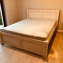 Jasna, drewniana rama łóżka z prostymi panelami, częściowo przykryta szarym prześcieradłem, stoi na drewnianej podłodze w pokoju z balkonem.