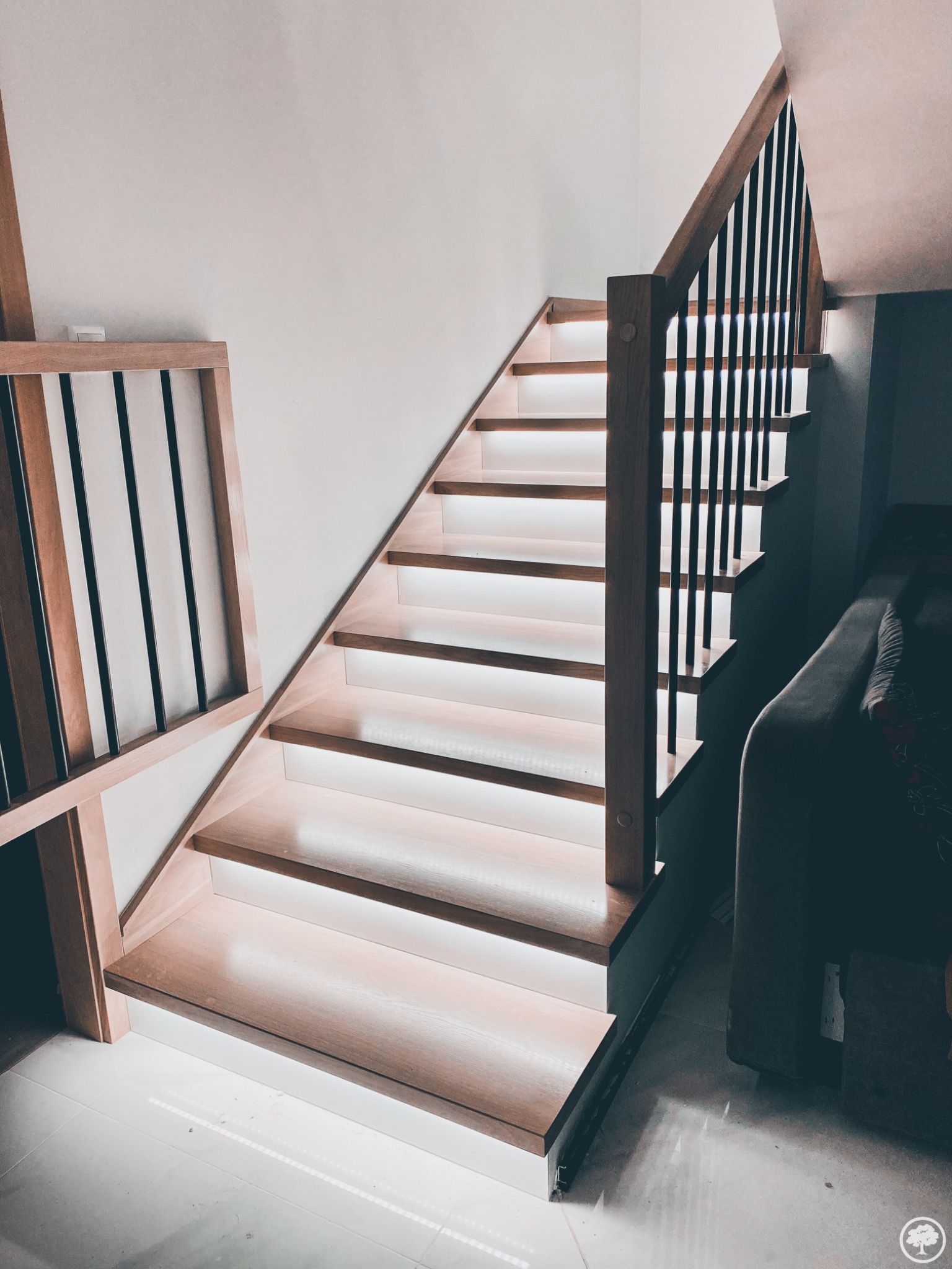 Drewniane schody z podświetleniem LED pod każdym stopniem, balustrada z drewnianymi słupkami i czarnymi, pionowymi elementami, widok od dołu, jasne płytki podłogowe.