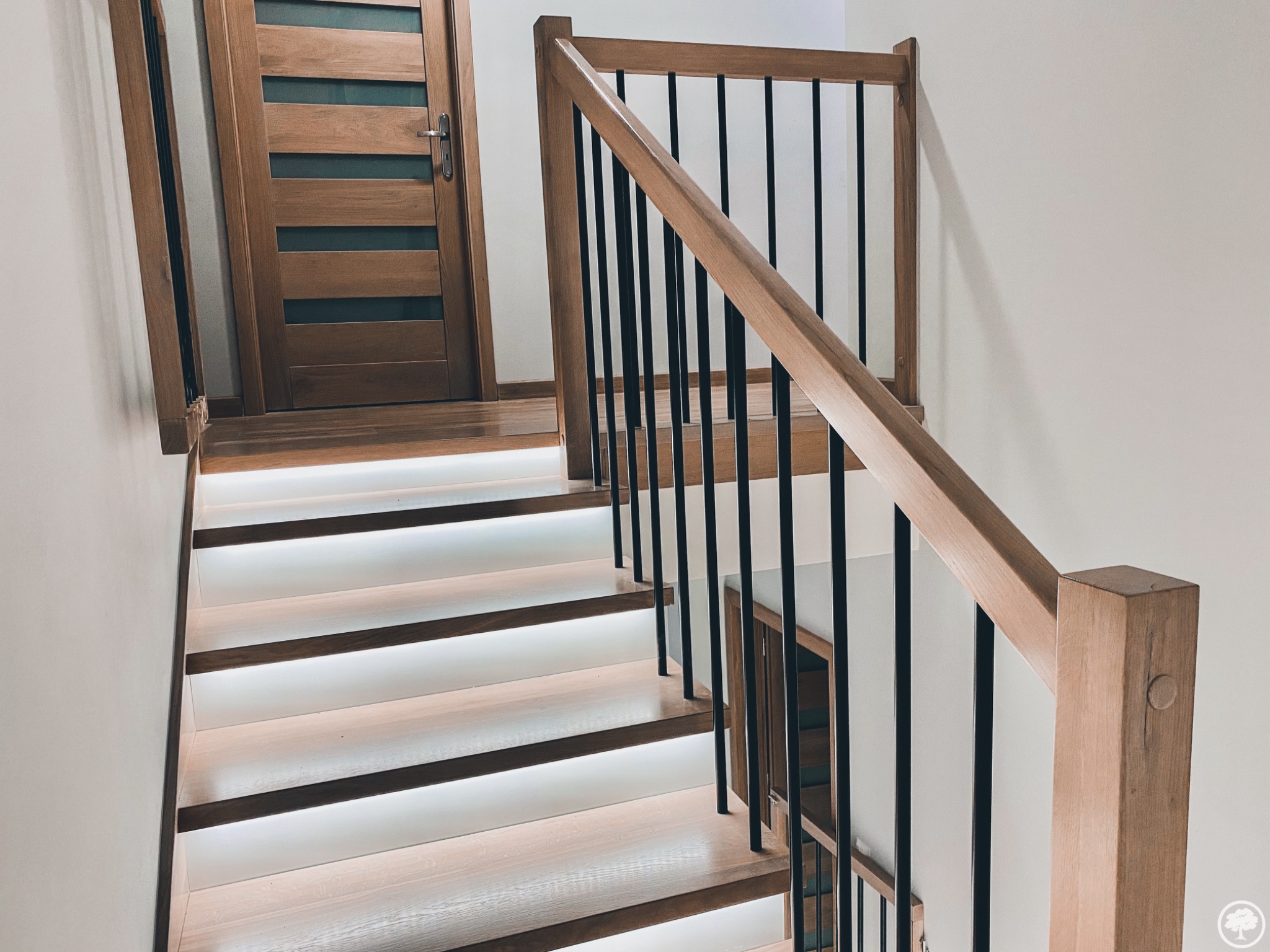 Nowoczesne schody drewniane z podświetlanymi stopniami i czarnymi balustradami, widoczne drzwi z drewnianą ramą i szybami.