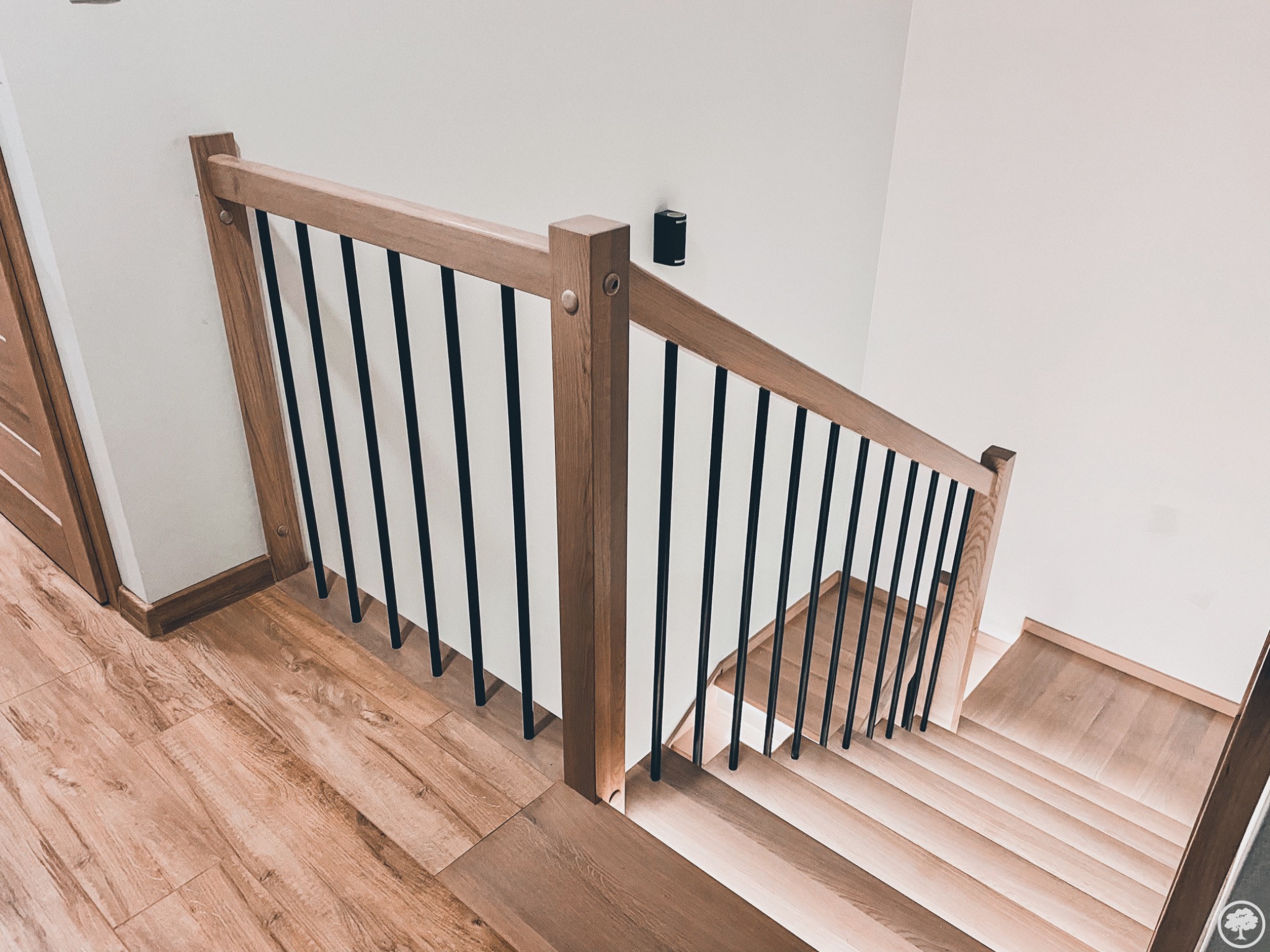 Widok z góry na drewniane schody z jasnego drewna z czarnymi metalowymi balustradami, oświetlenie ścienne w tle, widoczne fragmenty drewnianej podłogi i drzwi.