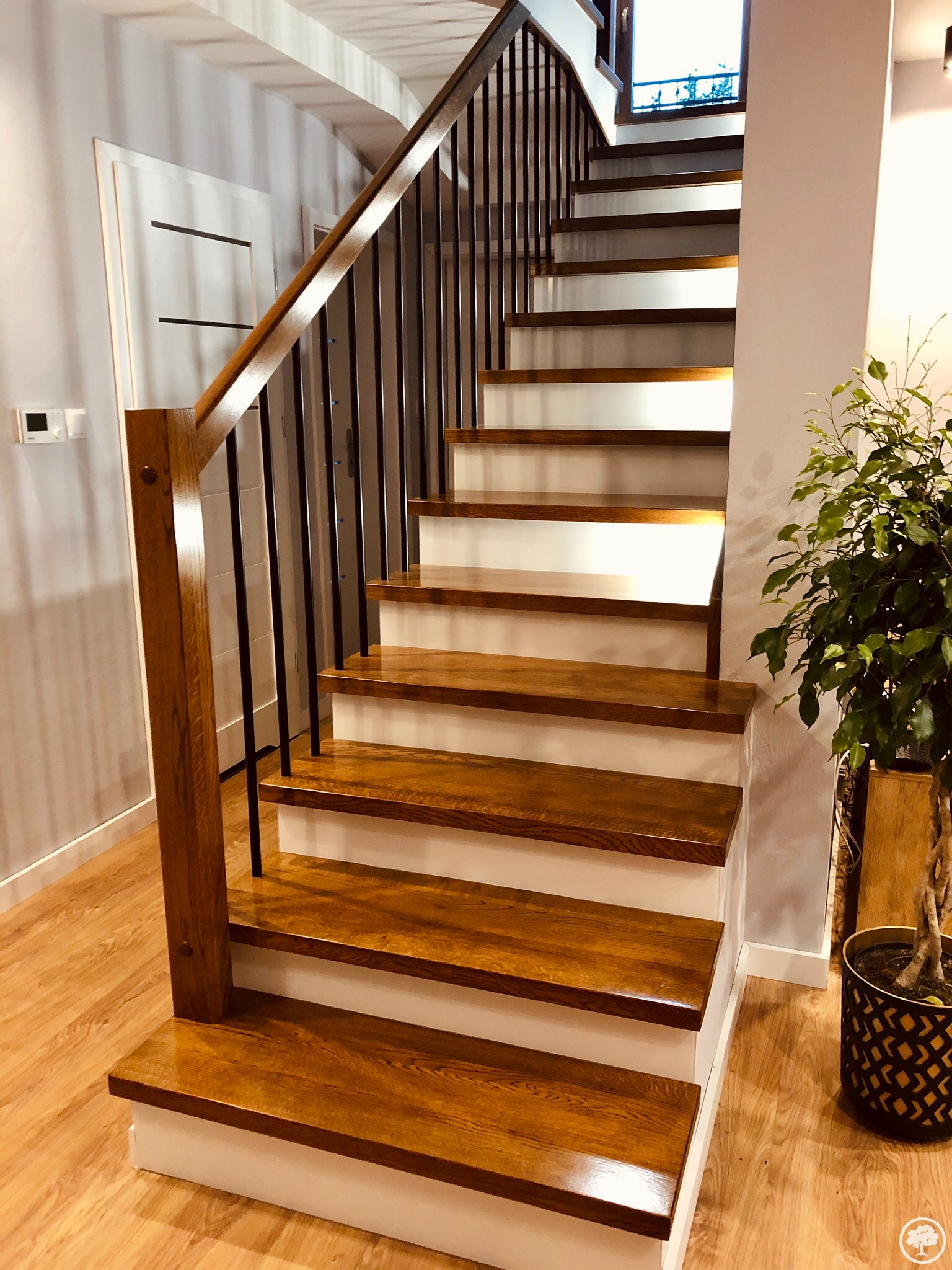 Nowoczesne schody drewniane z ciemnymi metalowymi balustradami i białymi podstopnicami, oświetlone ciepłym światłem, z widoczną rośliną doniczkową w tle.