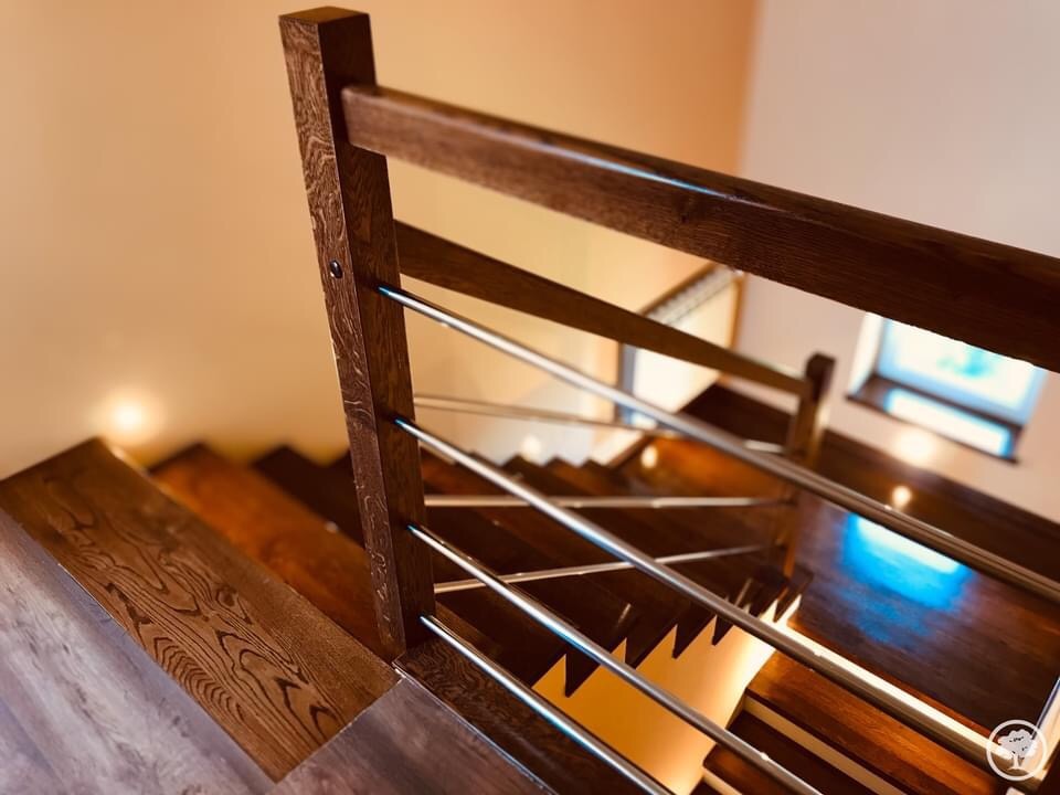 Klatka schodowa z drewnianymi schodami i balustradą z ciemnego drewna oraz metalowymi prętami, widok z góry w dół, oświetlenie punktowe.