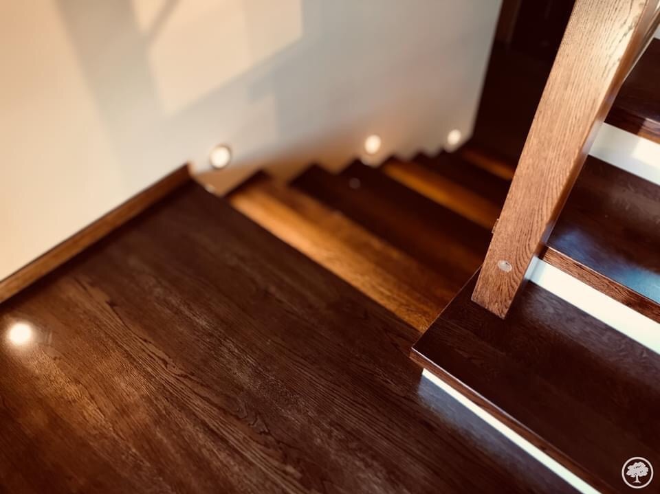 Nowoczesne schody z ciemnego drewna z podświetleniem punktowym wzdłuż ściany, widok z góry na stopnie i poręcz.