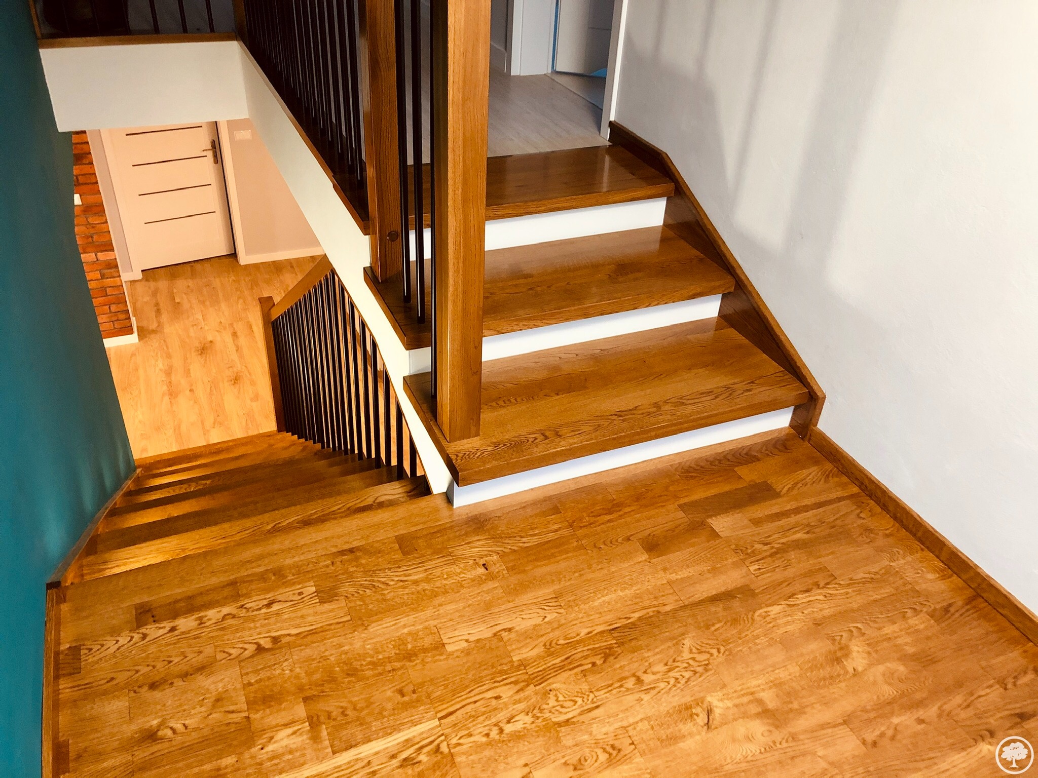 Widok z góry na drewniane schody z ciemnymi balustradami i białymi podstopnicami, prowadzące na drewnianą podłogę, widoczna ściana w kolorze turkusowym i białe drzwi.