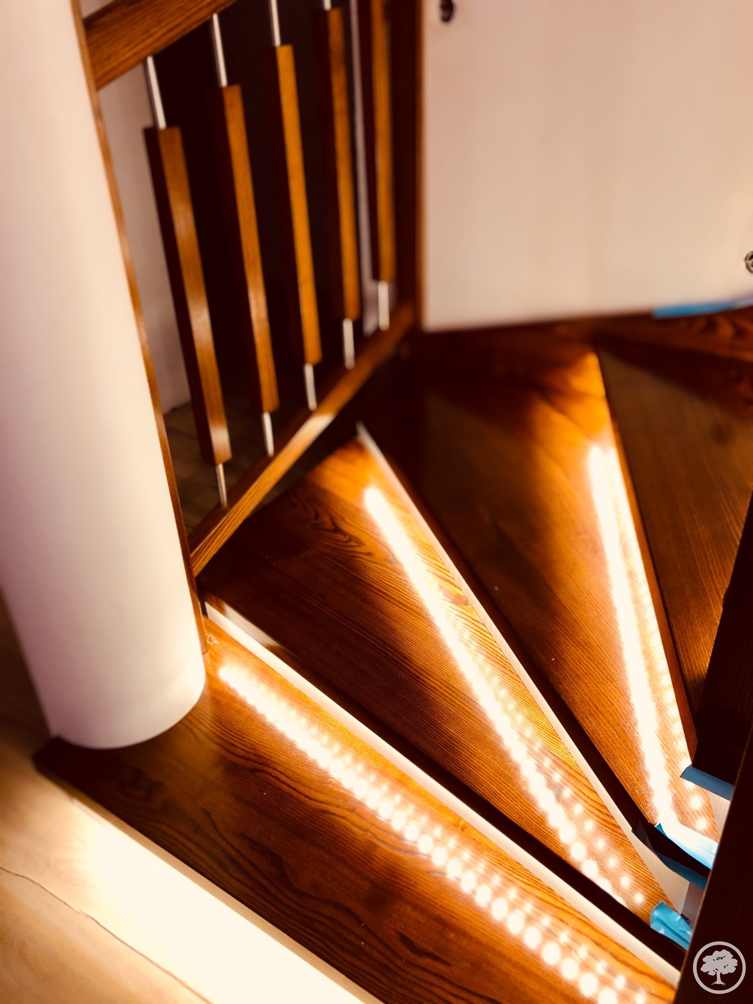 Wewnętrzne schody spiralne z drewna z oświetleniem LED pod stopniami, widok z góry, balustrada z drewnianymi szczebelkami i białym słupem.