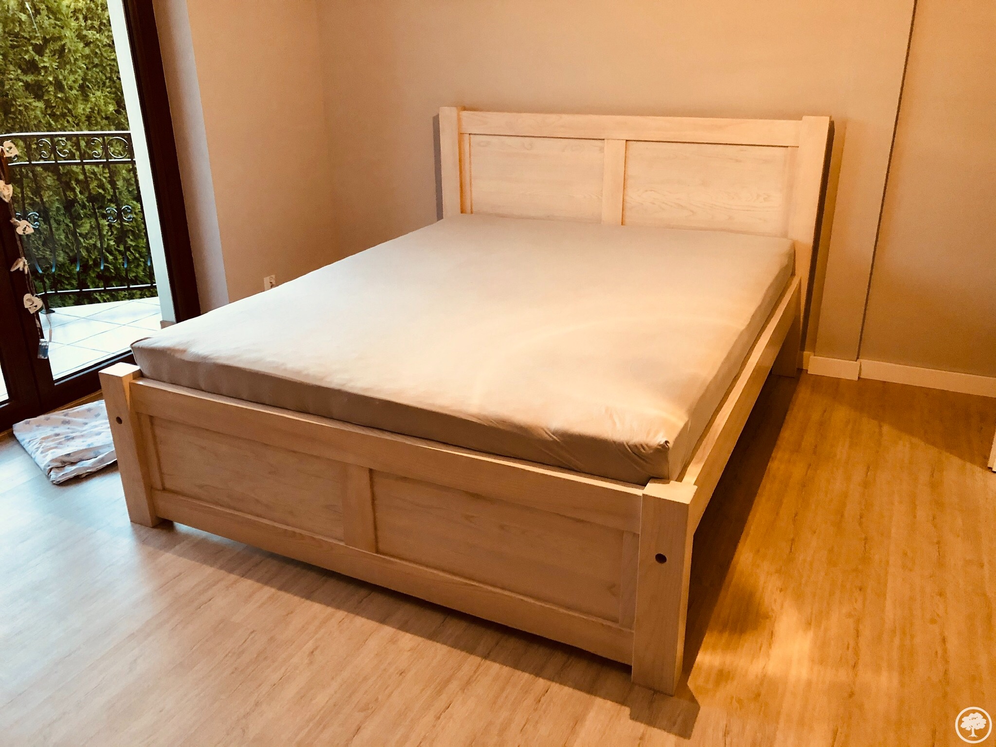 Jasna, drewniana rama łóżka z prostymi panelami, częściowo przykryta szarym prześcieradłem, stoi na drewnianej podłodze w pokoju z balkonem.